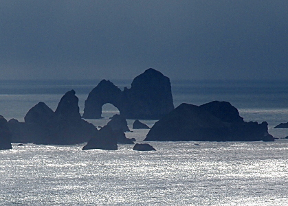 [Looking into the ocean one can sea many sea stacks and one open-arched rock. Because of the clouds, the rocks look dark while much of the foreground water has a white cast from the sun.]